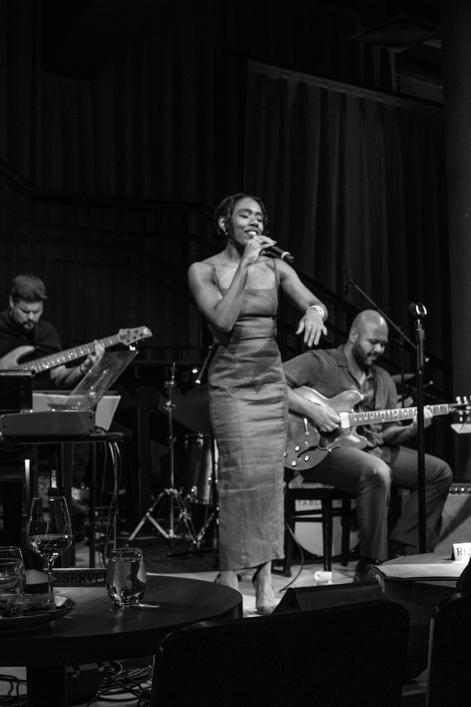Alicia Waller singing into a microphone with a bassist and guitarist behind her, photographed in black and white.