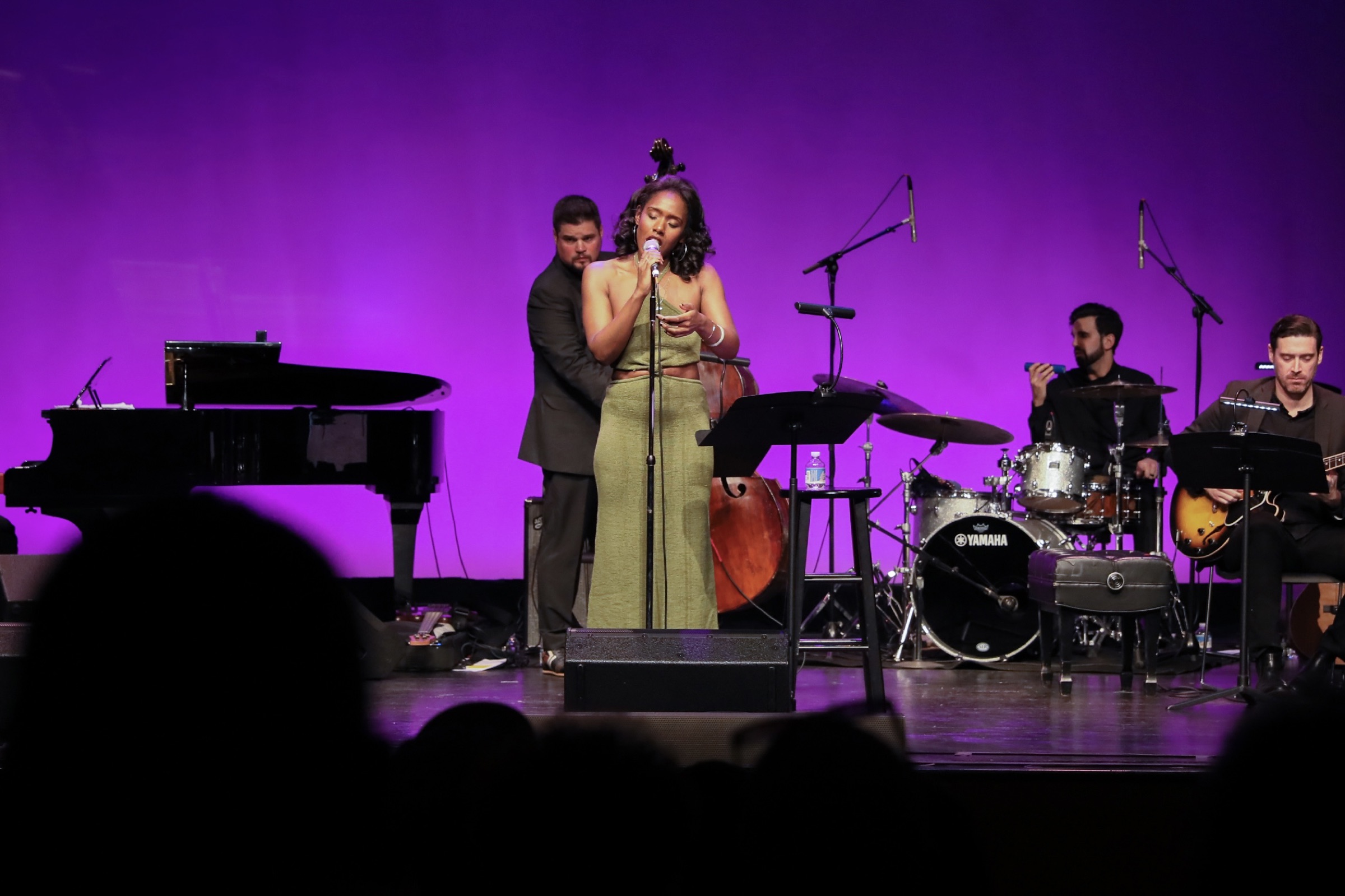 Alicia Waller performing with her full band, grand piano at stage left, under purple wash.