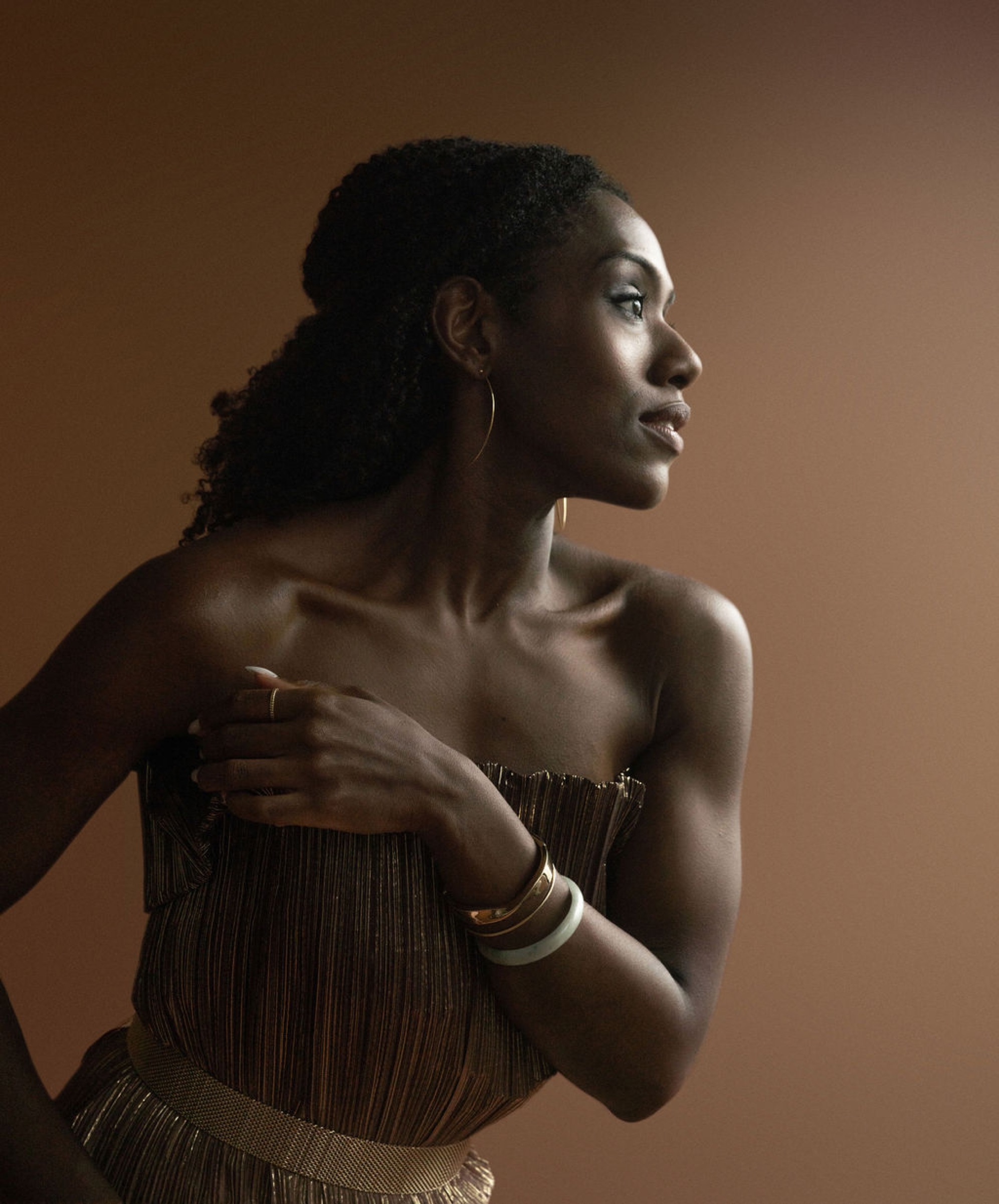 Alicia Waller in a gold pleated dress, hand at her collarbone, looking away from camera.