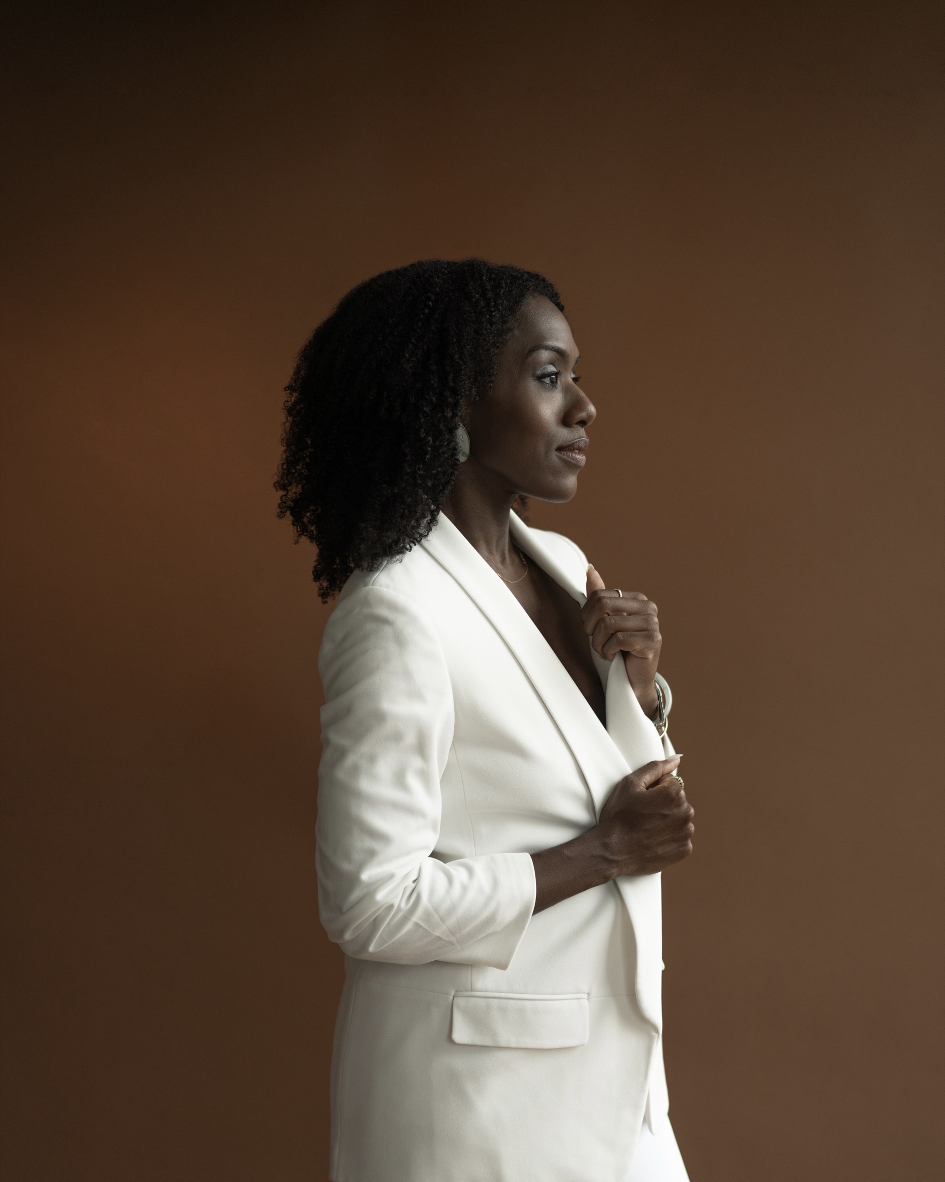 Alicia Waller in profile wearing a cream suit against a deep brown backdrop.